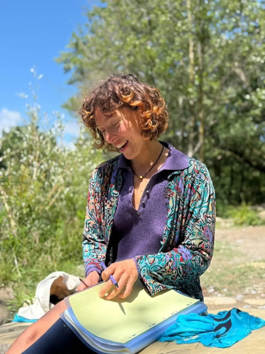 Pauline, souriante, assise au bord d'une rivière, carnet sur les genoux